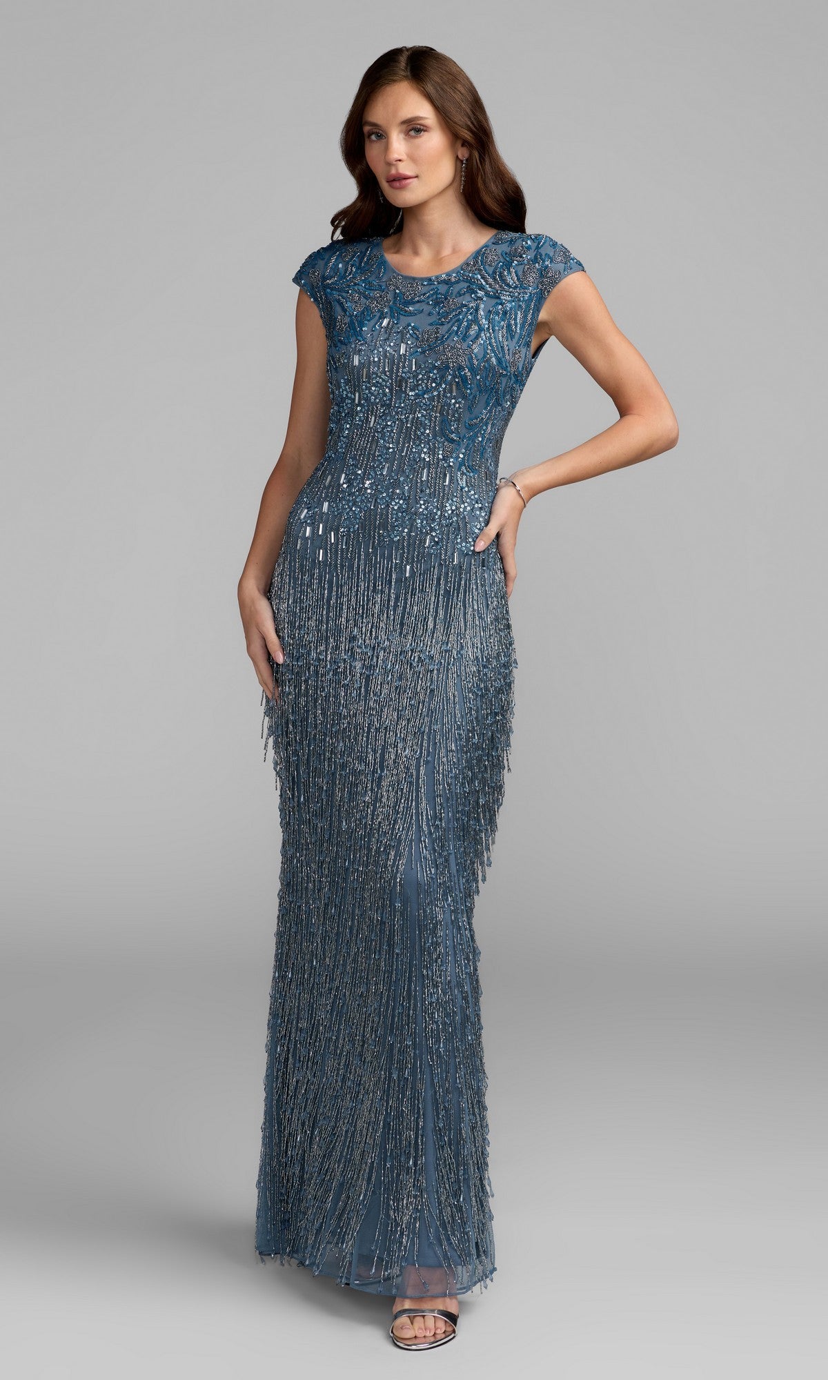 A woman in the Mac Duggal 6169 Beaded Long Lilac Formal Dress, featuring cap sleeves and shimmering beads and sequins, stands against a plain gray background.